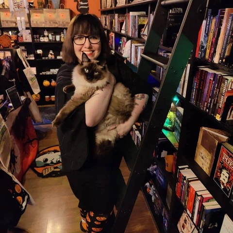 Death By TBR Books owner, Stephanie Csaszar poses in front of a bookshelf with her cat.